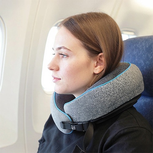 Woman sitting on an airplane wearing a gray travel neck pillow with a buckle, looking out the window.